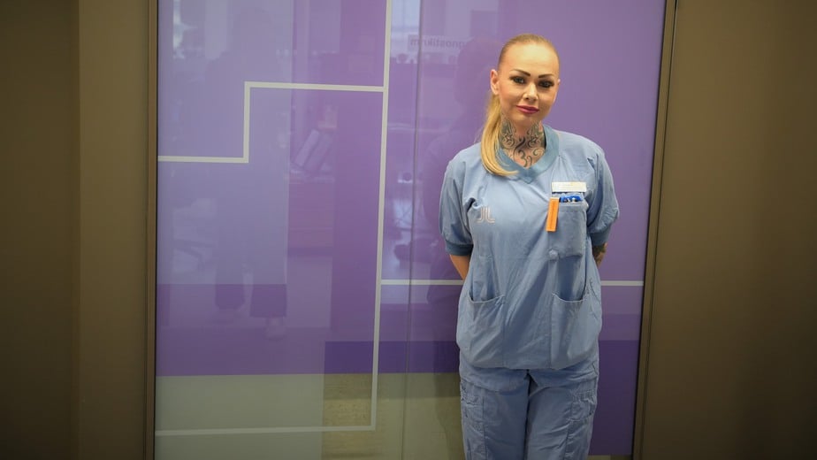 Linda Norin i blå sjukhuskläder står framför en rosa glasvägg