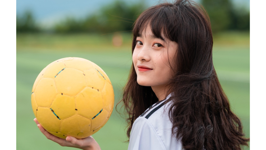 Tonårig tjej i vita fotbollskläder och en gul fotboll i handen.