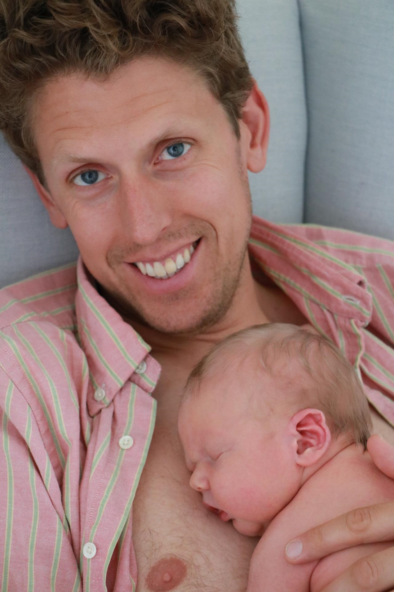 En man tittar leendes in i kameran, ett nyfött barn sover i hans famn