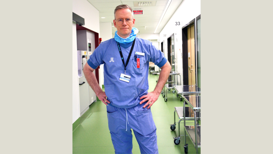 Olof Akre har blå operationskläder på sig och står in en sjukhuskorridor med grönt golv.